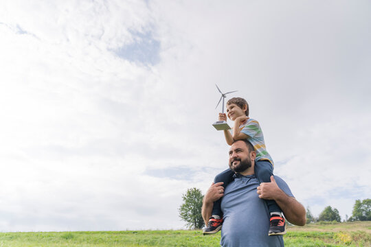 Man carrying son on shoulders with wind turbine model in front of cloudy sky