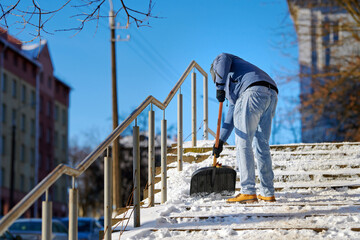 Man with snow shovel clear snow from the steps after blizzard, snow removal work. Worker with snowshovel in hands clean snowy staircase and walkway after winter storm