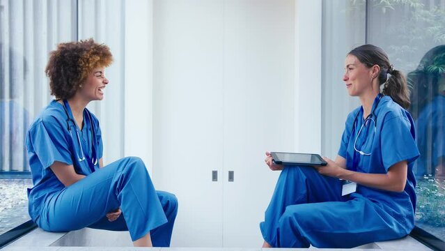 Two female doctors wearing scrubs with digital tablet taking a break talking together in hospital - shot in slow motion