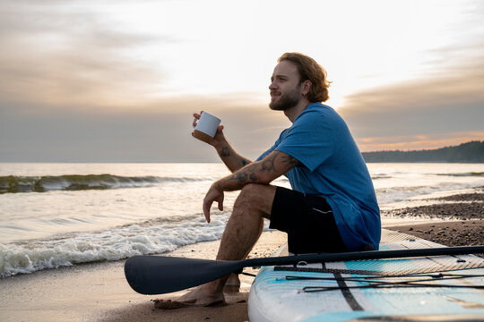 Young man having coffee sitting on paddleboard at beach