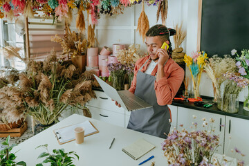 Young entrepreneur talking on smart phone and using laptop in flower shop