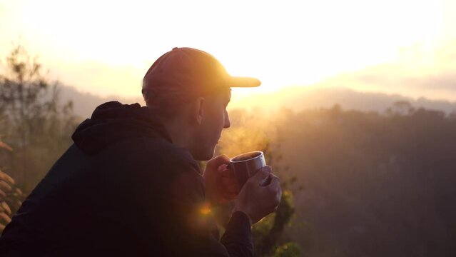 Millennial man in hoodie enjoying cup of coffee on warm sunrise in mountains. Cinematic side view of stunning sunrise with sun rays through morning fog and traveler in cap. Meet dawn early in sunrise