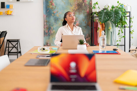 Young female boss sitting at wooden table with laptop and working in modern workspace during daytime while looking away