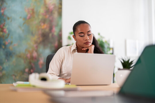 Serious female manager in white shirt sitting at table with laptop and looking at screen in light contemporary office