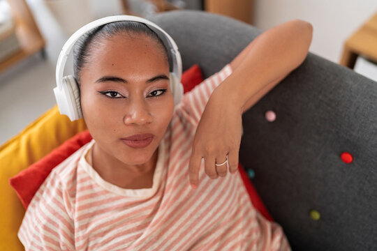 High angle of young Asian female in striped t shirt listening to good music on headphones while resting on comfortable couch and looking at camera