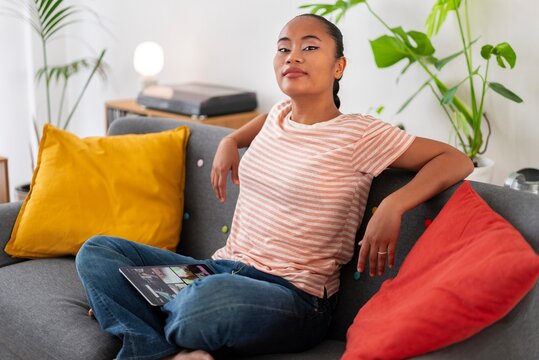 Young Asian female artist sitting on sofa with crossed legs wearing striped t shirt with tablet during free time