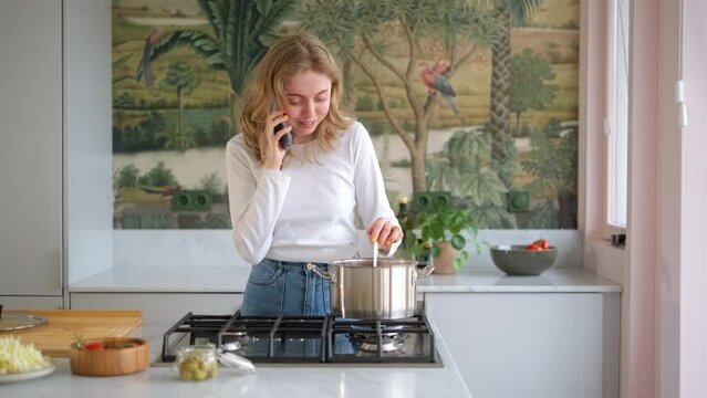 Cute young woman holds cellphone talks on phone while cooking healthy vegetarian soup in domestic kitchen. Сover the pot. Pleasant distancing communication food delivery services by phone call cookery