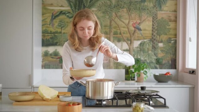 Young beautiful woman pours hot soup into bowls with ladle in cozy kitchen. Domestic food, cooking at home, housewife doing chores