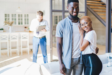 Happy ethnic couple hugging while standing in new apartment with estate agent