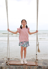 Portrait of Asian young girl play standing on swing against cement wall at seaside. Full length.