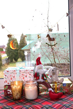 toy gnome with teddy bear sitting on christmas gifts with candles near window with garland in rural house, vertical photo