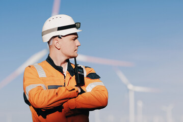 Wind Farm Offshore Maintenance Technician. Seafarer. Seaman. Navigator. A Man In A Working Overall Boiler Suit With A Radio And Safety Helmet With A Blurred Wing Generators In The Background - obrazy, fototapety, plakaty