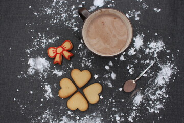 Black tablecloth full of flour, after having made homemade sweets for Christmas celebration. A mug of hot chocolate or coffee, heart gingerbread and lace cookies and a spoon with cocoa. Winter scene.