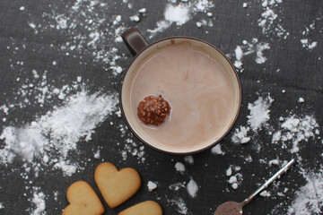 Black tablecloth full of flour, after having made homemade sweets for Christmas celebration. A mug of hot chocolate with a floating little round chocolate, heart cookies and a spoon with cocoa.