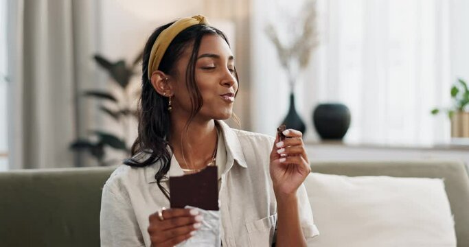 Woman, eat and chocolate to enjoy in home with bite for candy, snack and delicious. Indian person, hungry and sugar for comfort in diet, nutrition or calories for cacao craving on sofa in living room