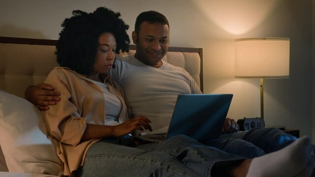 Happy family African American married couple husband man and wife woman on bed at night evening browsing net laptop using computer talking shopping app in internet choose film movie in online service