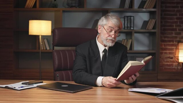 Attentive aged male in formal clothes and reading glasses looking at book page while staying in upscale workplace. Confident general manager getting advice on business strategies in comfort of office.