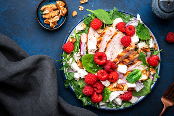 Tasty salad with raspberries, grilled chicken, cheese, onion, walnuts, spinach and arugula, blue table background, top view