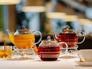 Set of three transparent glass teapots with warmer and candles, hot herbal tea on table on restaurant bokeh background. Mango tea, rosehip herbal tea, spicy herbal tea with star anise and honey - tea
