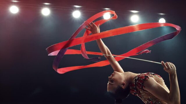 Portrait of a Graceful Rhythmic Gymnast Performing a Dance with a Red Ribbon. Young Female Exercising in a Striking Beautiful Uniform in a Dark Studio. Cinematic Super Slow Motion Speed Ramp Footage