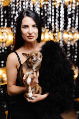 Young beautiful smiling brunette female in trendy evening clothes holding her litlle dog in her hands. Carefree woman posing near shiny tinsel wall in studio with dog.