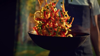 Chef Stir Frying Vegetables in a Flaming Wok in an Outdoors Kitchen. Person Cooking Food Using a Flambe Technique, Igniting Hot Oil and Alcohol in a Pan. Close Up Super Slow Motion Footage