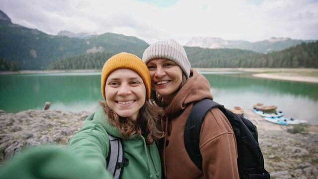 A romantic lesbian couple takes a selfie and kisses against the background of a lake while hiking in the mountains.
