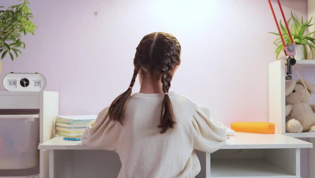 Tired exhausted little girl wearing white sweater with pigtails sitting at table doing home work felling pain in neck massaging painful back.