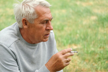 Senior man lighting up a cigarette outdoors against green nature background. Old thoughtful man smoking, looking aside. Close Up Face. Relaxed pensioner enjoy weekend rest outside. Unhealthy habits