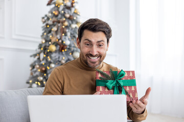 A young happy man is sitting at home on the sofa near the Christmas tree and talking on a video call on a laptop, congratulating on the New Year and winter holidays, holding a gift in his hands