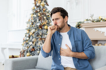 Portrait of a young man who fell ill during the festive Christmas holidays and weekend. Sitting at home on the sofa and coughing, covering his mouth with his hand and holding his chest