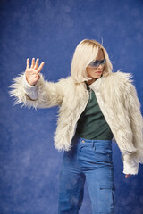 young woman in trendy sunglasses and white faux fur jacket gesturing under falling snow on blue