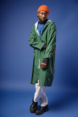 full length, bearded african american model in knitted hat and winter attire posing on blue backdrop