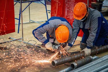 Craftmans grind steel pipes. Men use hand-held circular saw. Processing pipes for scaffolding construction. Craftmans are sawing off elements for scaffolding. Craftmans polish pipeline - obrazy, fototapety, plakaty