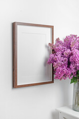 Blank wooden picture frame mockup with fresh lilac flowers bouquet over white wall, frame mock up with copy space