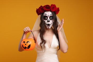 Emotional young woman in scary bride costume with sugar skull makeup, flower crown and pumpkin bucket on orange background. Halloween celebration