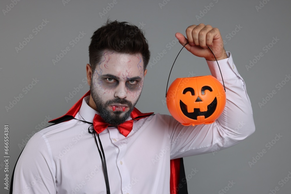 Poster man in scary vampire costume with fangs and pumpkin bucket on light grey background. halloween celeb