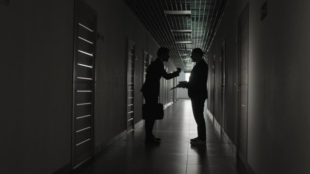 Full shot of two male figures in formal suits arguing while standing in dark long hallway of office building