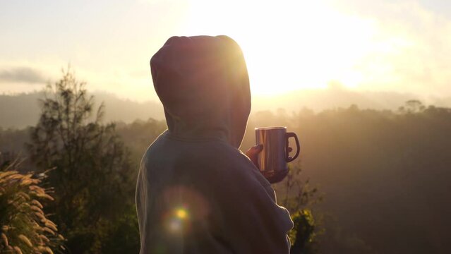 Woman in hoodie enjoying cup of coffee on warm sunrise in mountains near camping van. Cinematic side view of stunning sunrise with sun rays through morning fog and traveler. Meet dawn early in sunrise