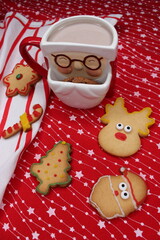 Fototapeta premium A Santa Claus-shaped mug filled with hot chocolate or coffee. A chocolate chip cookie peeks out of the cup. Homemade Christmas cookies. Red tablecloth with white stars and a striped kitchen towel. 