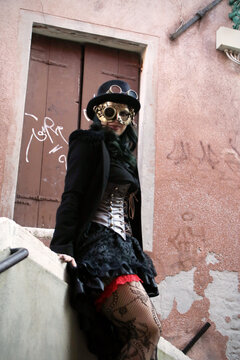 Steampunk girl in stockings, dress, corset, top hat, and retrofuturistic glasses poses in Venice canals. Her stylish attire and charming charisma captivate.