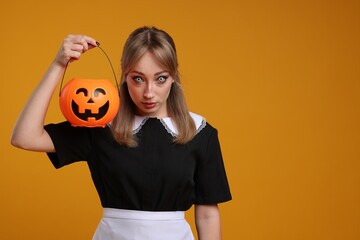 Fototapeta na wymiar Woman in scary maid costume with pumpkin bucket on orange background, space for text. Halloween celebration