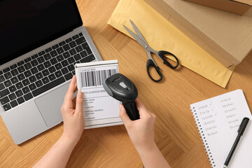 Woman with scanner reading parcel barcode at wooden table, top view. Online store