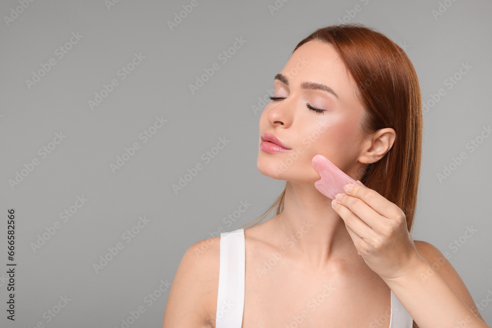 Sticker Young woman massaging her face with rose quartz gua sha tool on grey background, space for text