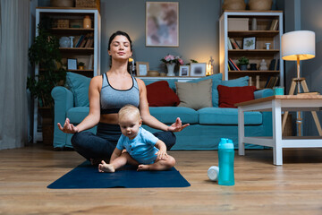 Young happy successful independent single mother is practicing yoga at home on a yoga mat while her baby is carelessly sitting on the floor and playing with a water bottle. Mom stretching muscles