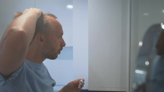 Man use anti baldness serum or product, dripping from pipette onto his bald patches in bathroom and looks at camera.