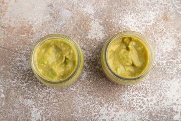 Baby puree with vegetable mix, broccoli, avocado in glass jar on brown concrete, top view
