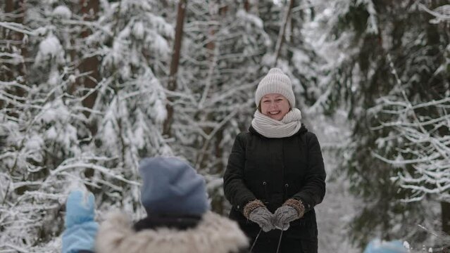 Portrait of happy nanny carries child on sled and cutely waves her hand in mitten to him in snowy forest. Woman and child walk in warm clothes through fabulous winter forest on vacation.
