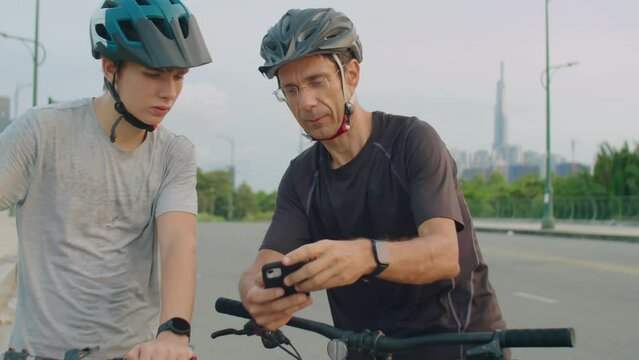 Dad and son in sportswear and helmets leaning on bikes, using map on mobile phone and discussing route while standing on road