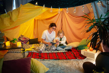 A higher camera shot of a modern and cozy looking indoor blanket tent where there is a wholesome moment of a mother and her son sharing a bonding time while reading together